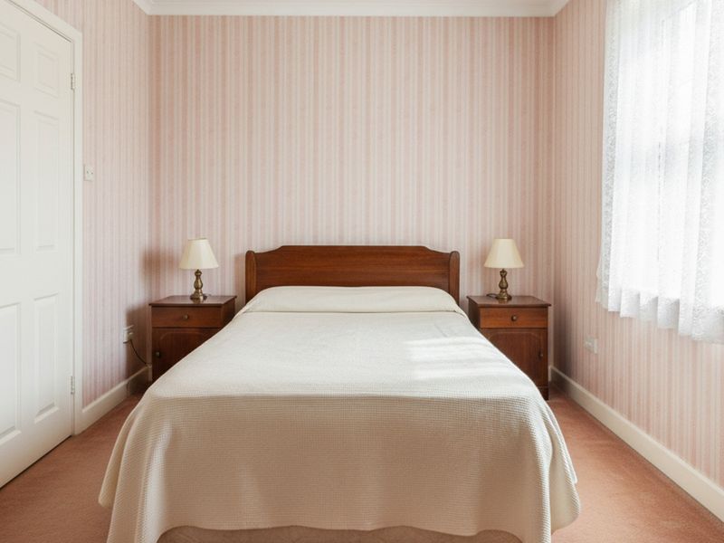 A small bedroom with pink striped wallpaper and pink carpet, a dark wooden bed frame, and two matching bedside tables with cream lampshades.